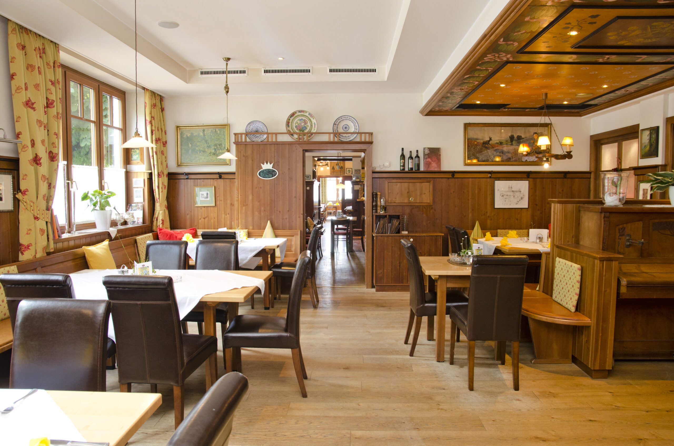 A bright and cozy dining room with large windows, warm wooden paneling, and neatly set tables. Dark leather chairs, traditional decor, and a painted wooden ceiling create an inviting atmosphere that blends tradition with comfort.
