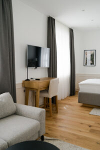 Modern hotel room with a TV, wooden desk, armchair, and bright natural light
