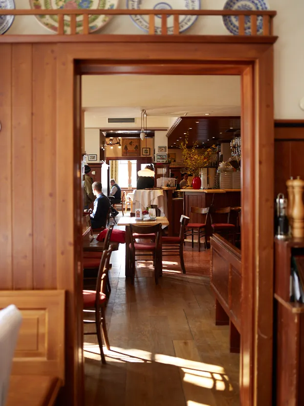 Blick durch einen Holzrahmen in den sonnigen Gastraum mit Tischen, Stühlen und Gästen.