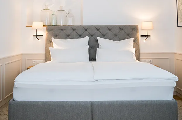 Modernes Hotelzimmer mit grauem Polsterbett, weißer Bettwäsche und warmen Wandleuchten.
