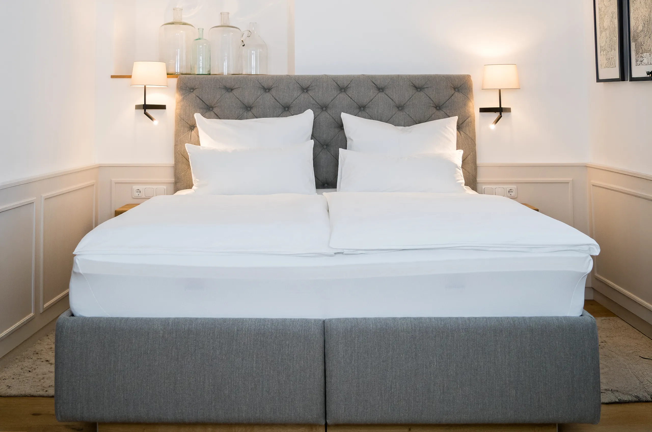 Doppelbett mit grauem, gestepptem Kopfteil und weißen Kissen in einem hellen Hotelzimmer.
