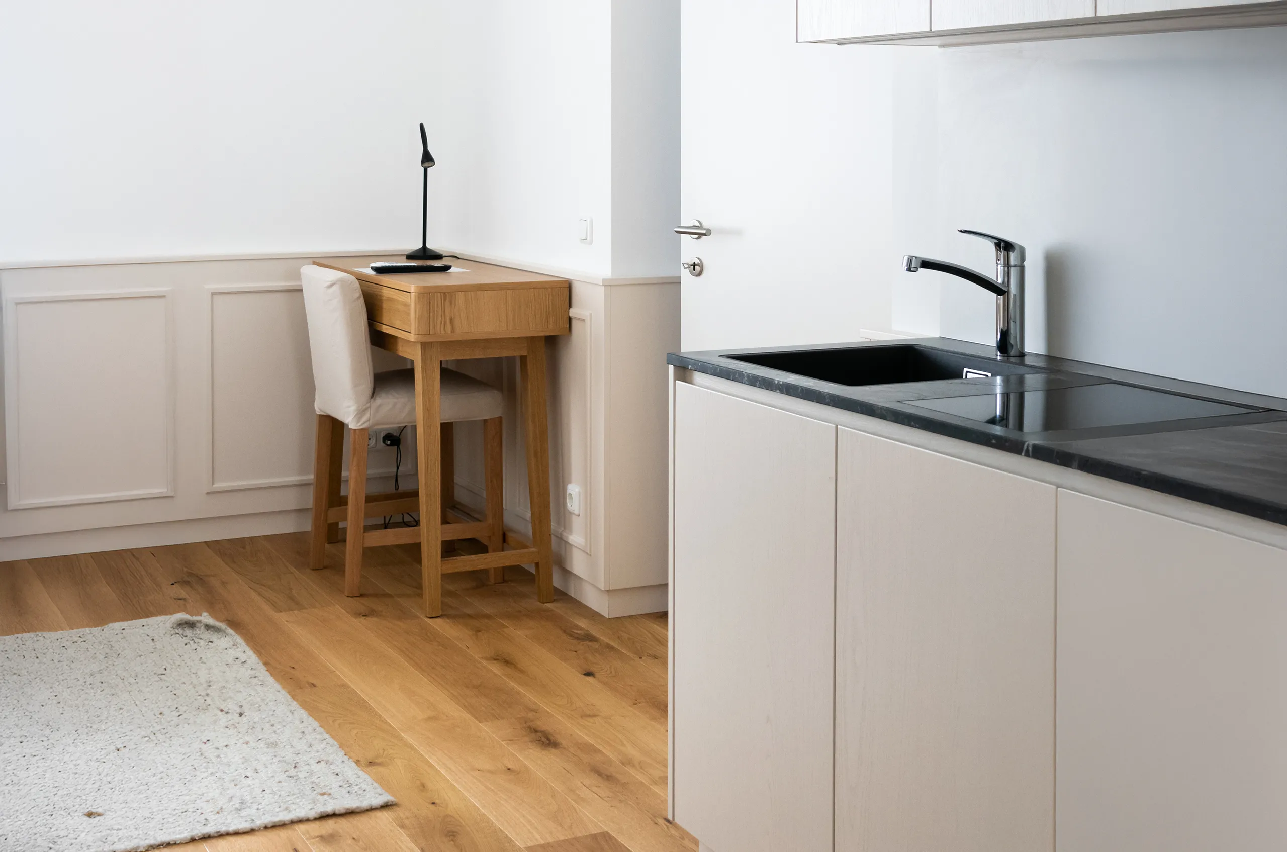 Arbeitsplatz mit hellem Holzschreibtisch, Stuhl und moderner Küchenzeile im Hintergrund.