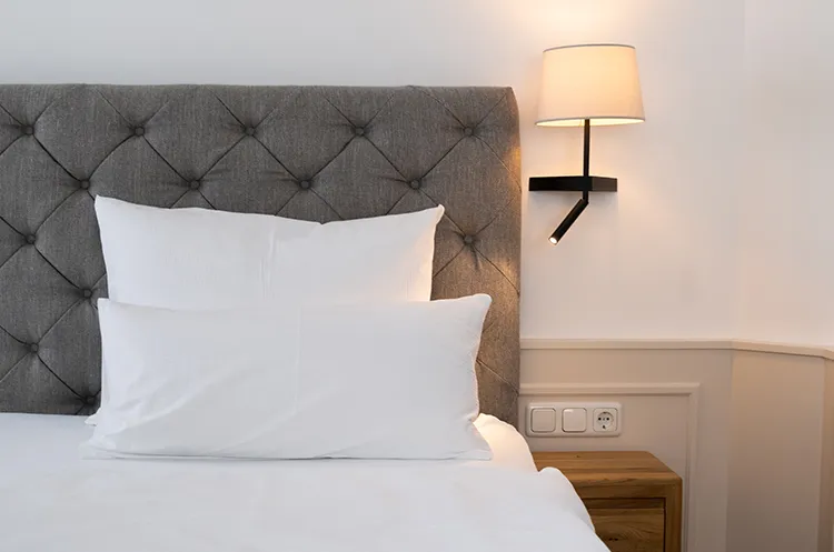 Nahaufnahme eines Betts mit grauem Kopfteil und weißen Kissen sowie Wandlampe.