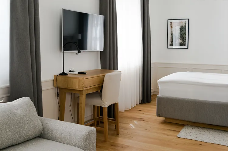 Helles Zimmer mit Schreibtisch, Stuhl, Fernseher und grauem Sofa im Vordergrund.