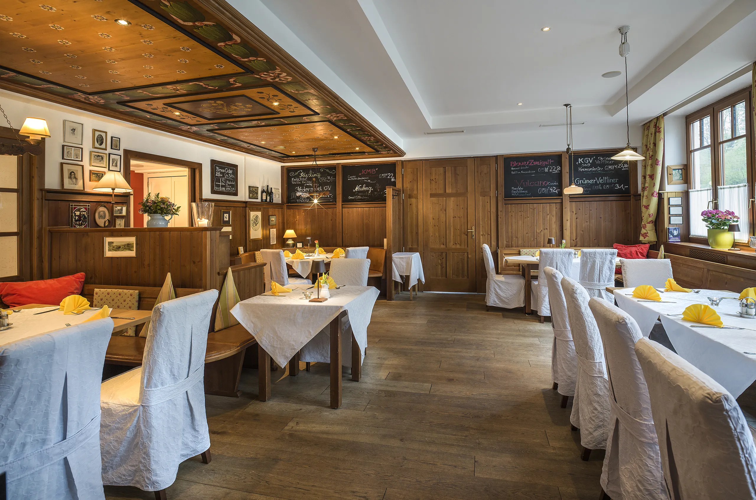 Ein gemütlicher, traditioneller Speiseraum mit Holzvertäfelung, bemalter Decke und liebevoll gedeckten Tischen mit weißen Tischdecken und gelben Servietten – eine warme und einladende Atmosphäre.
