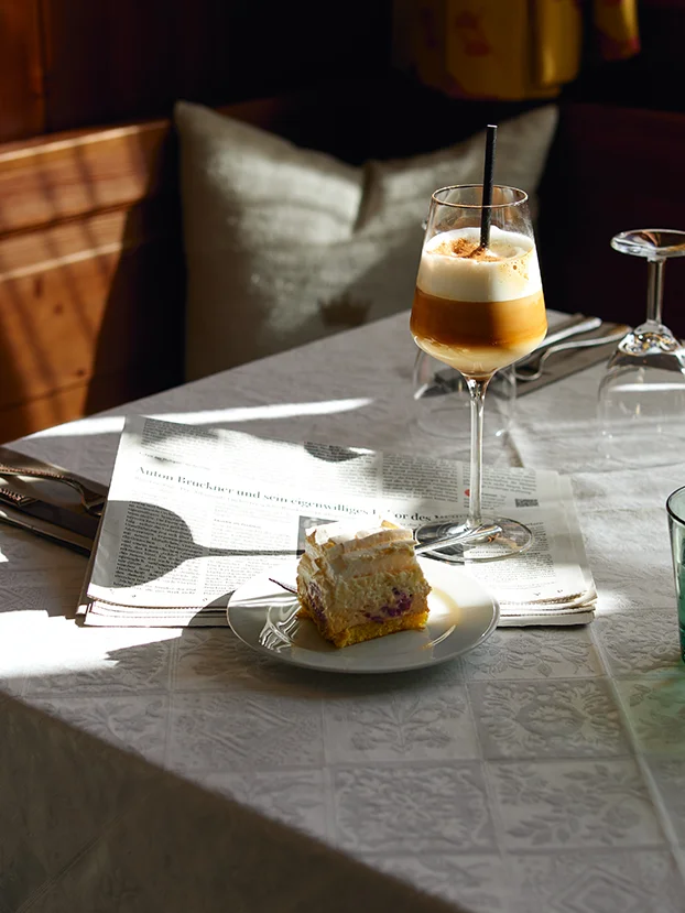 Ein Stück Cremetorte und ein Latte macchiato auf einem sonnigen Frühstückstisch mit Zeitung.