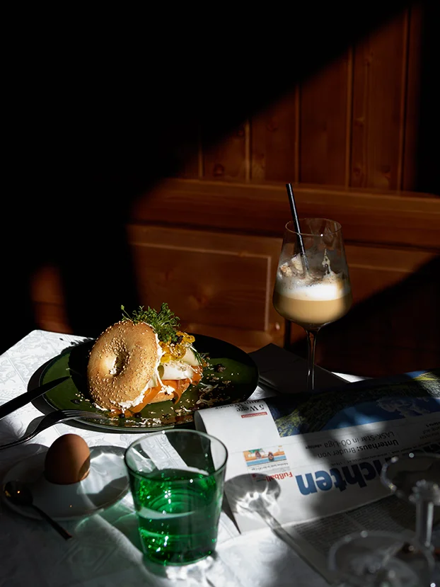 Frühstückstisch im Sonnenlicht mit belegtem Bagel, einem Glas Kaffee, grünem Wasserglas und Zeitung in gemütlicher Holzatmosphäre.